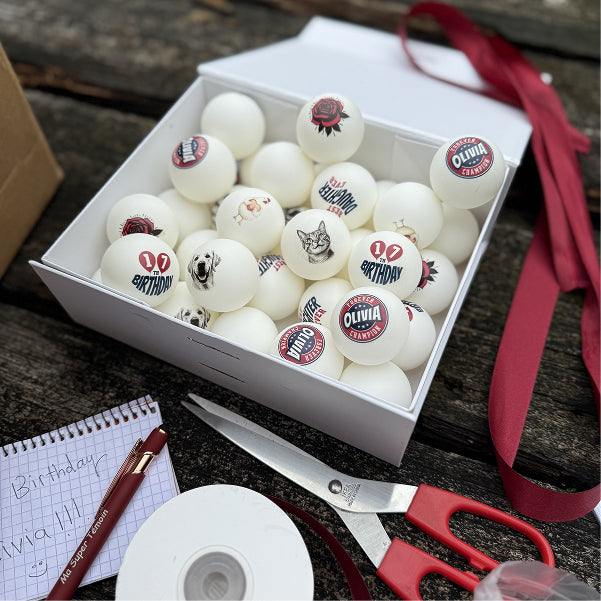Box filled with personalized ping pong balls with different designs on a wooden surface with scissors and ribbon.