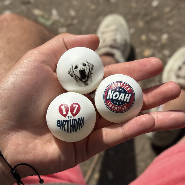 Three personalized ping pong balls held in a hand with a blurred background