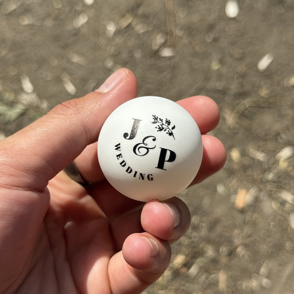Hand holding a white ball with 'J & P Wedding' text against a blurred natural background
